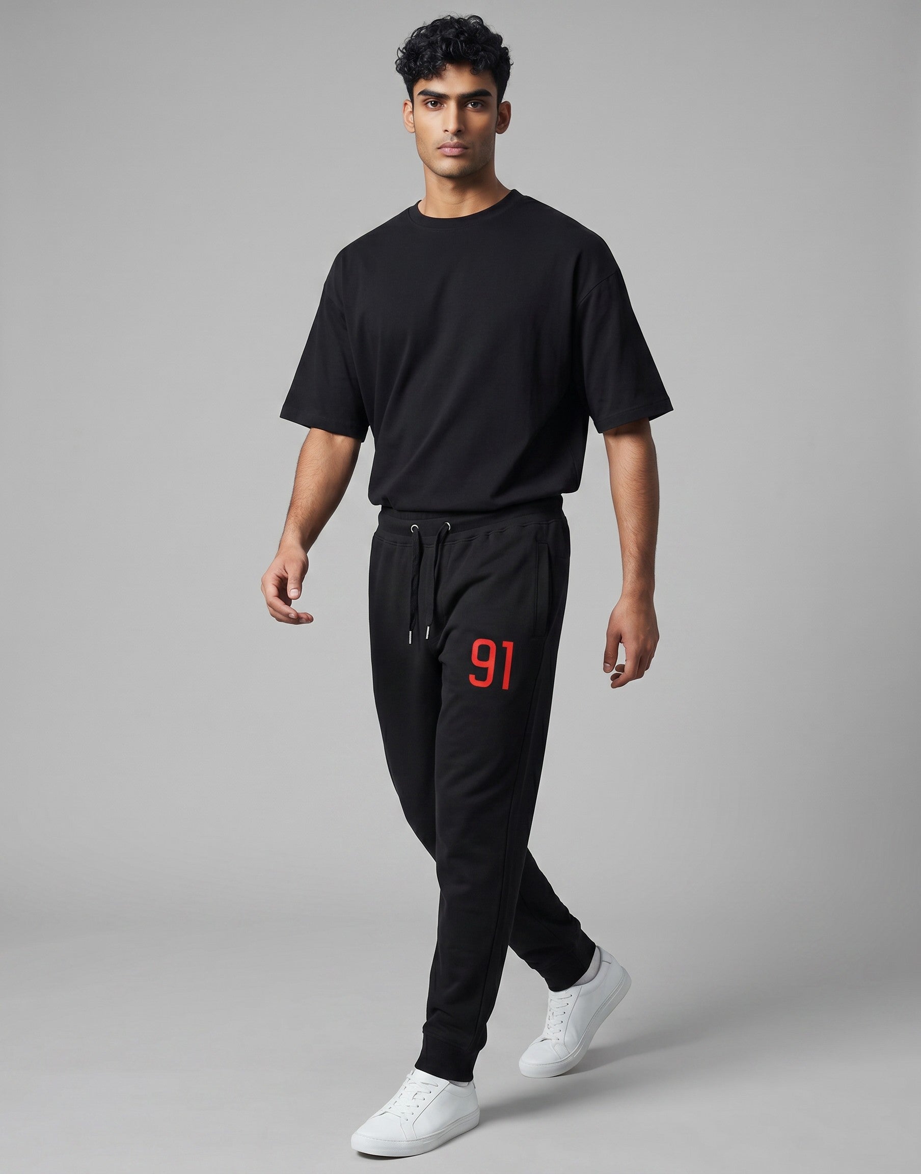 A male model wearing Tribe Neo 91 black cotton fleece joggers with a red 91 graphic on the thigh and white sneakers.