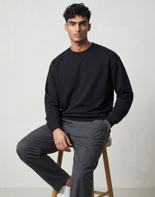 Tribe Neo 91 black oversized unisex sweatshirt with dropped shoulders worn by a man sitting on a wooden stool