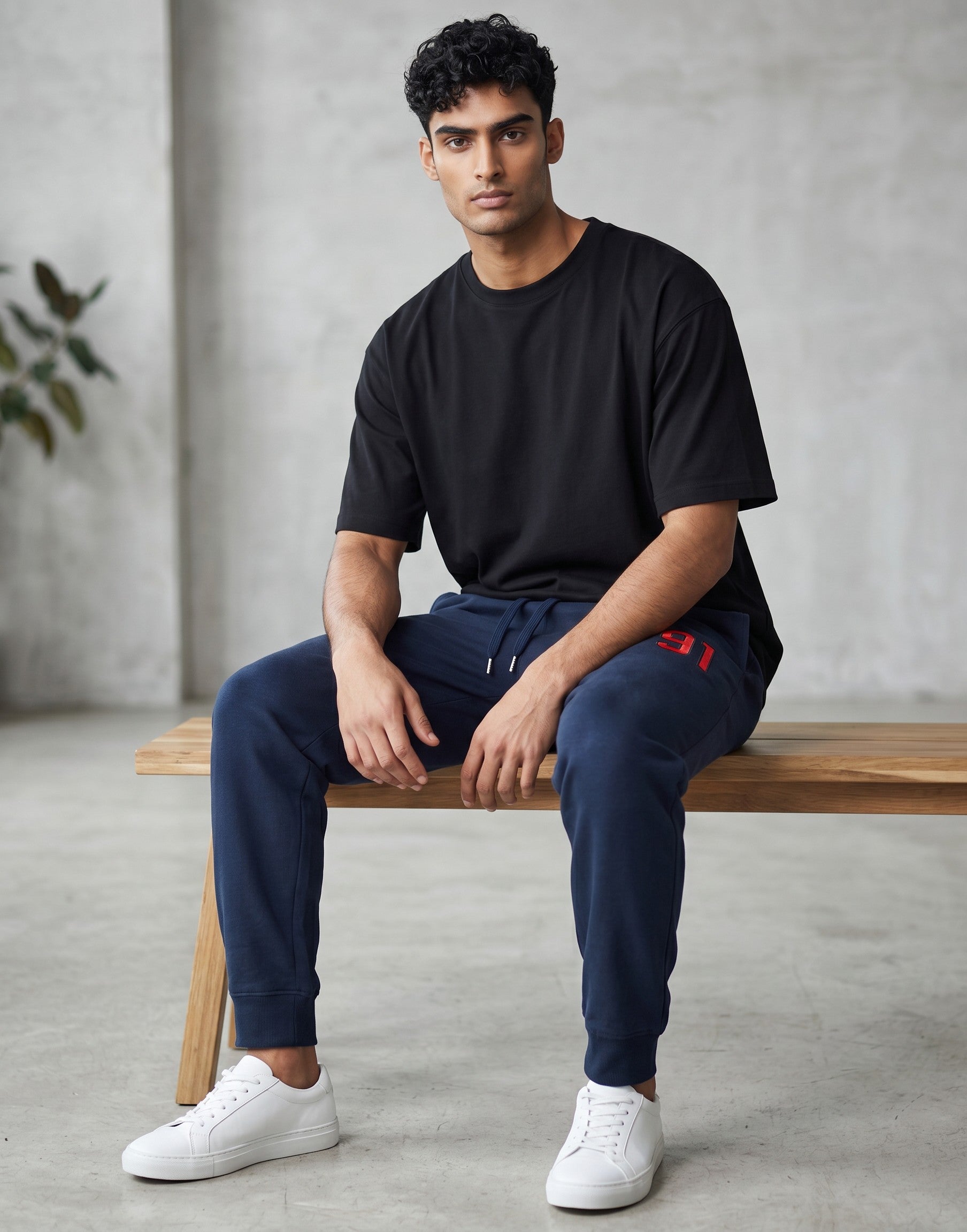 A man wearing Tribe Neo 91 navy blue cotton fleece joggers with a red 91 graphic, sitting on a wooden bench.